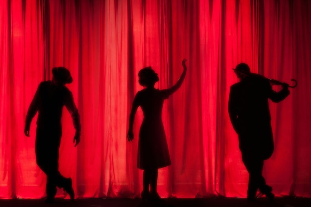 Silhouetted actors in dramatic poses on stage, with a red curtain backdrop