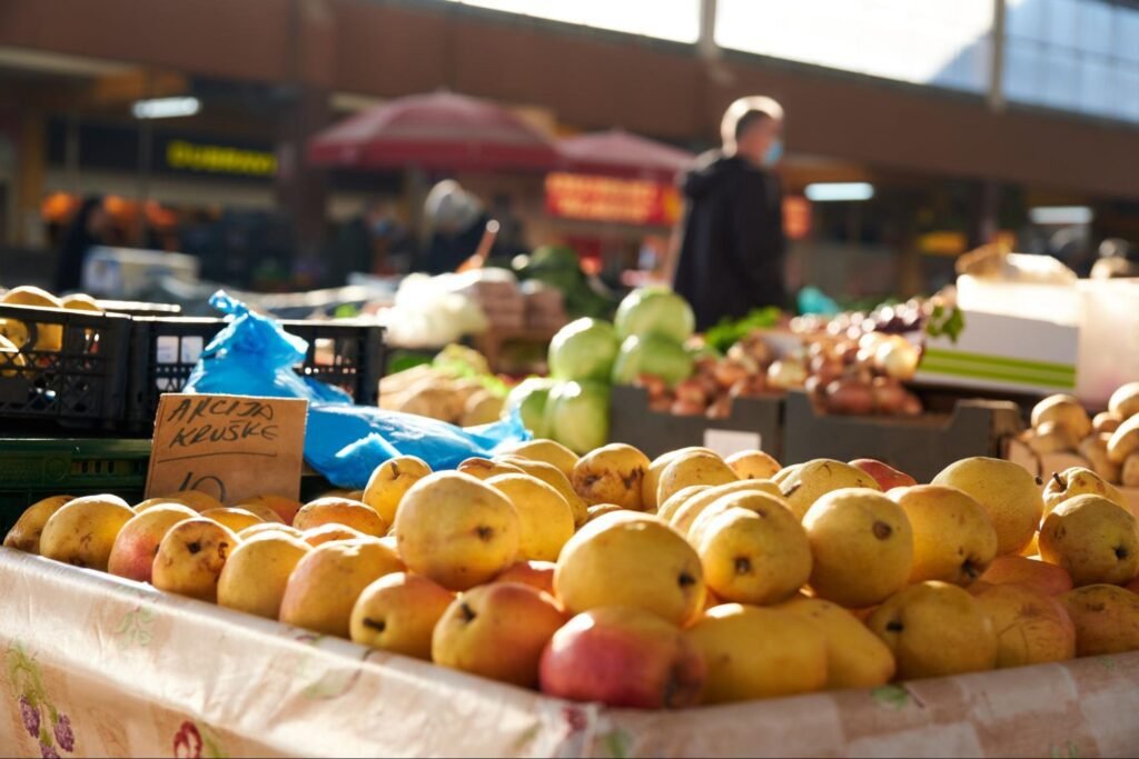 Fresh produce displayed at an outdoor farmers market with a vendor and shoppers in the background