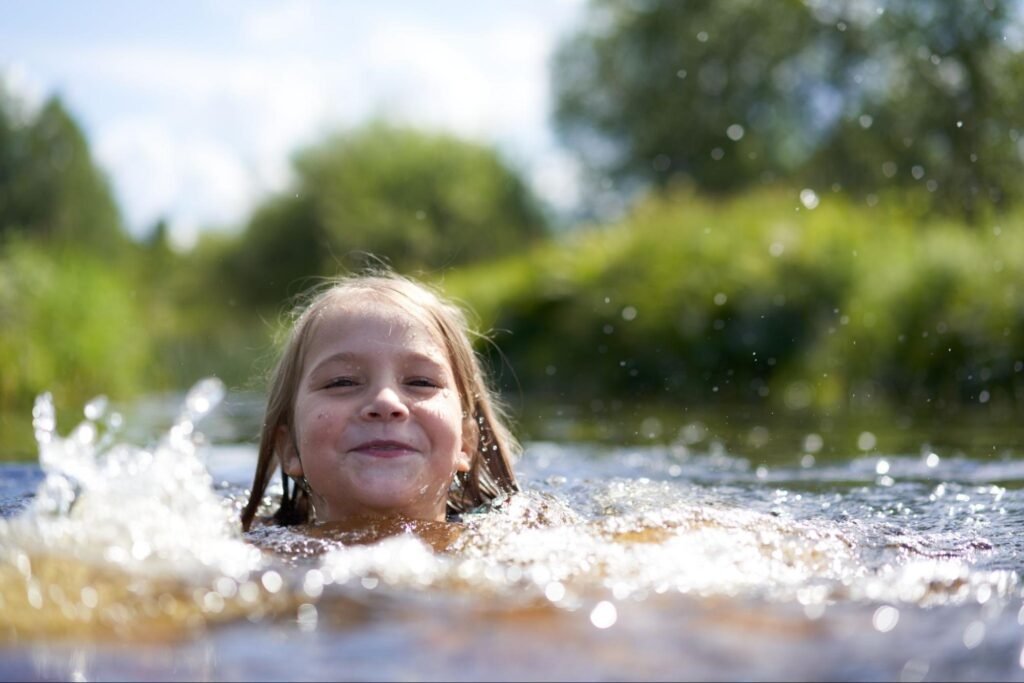 A young child smiling while swimming in a calm lake surrounded by greenery.